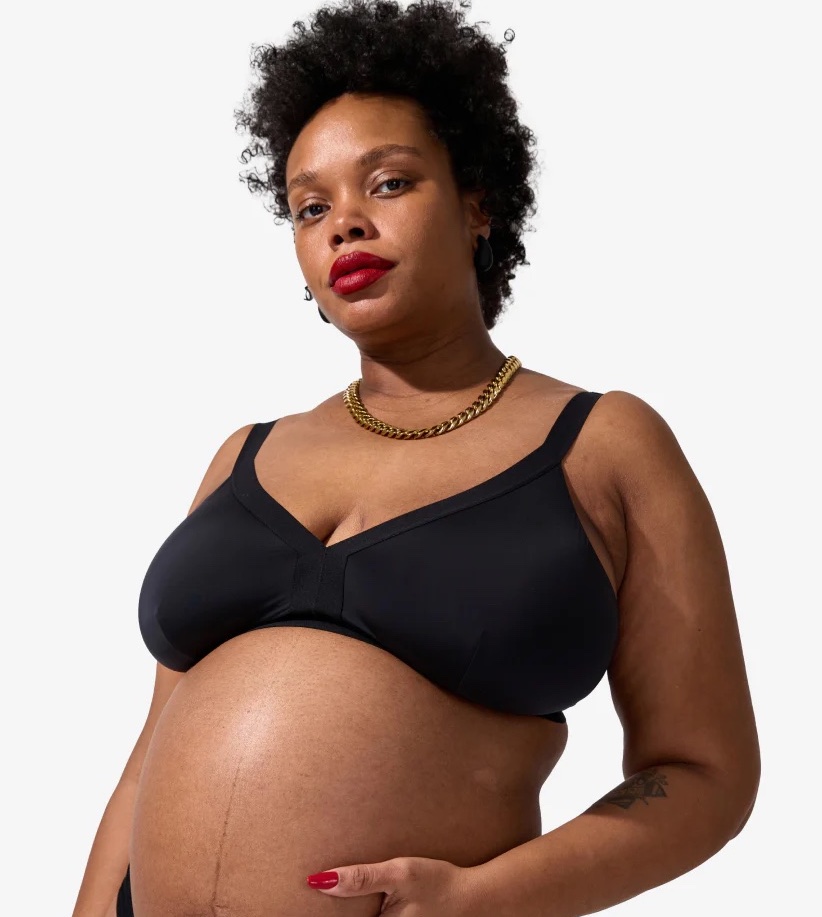 A pregnant person wearing a black bra and gold chain necklace stands against a plain background, displaying a visible baby bump.