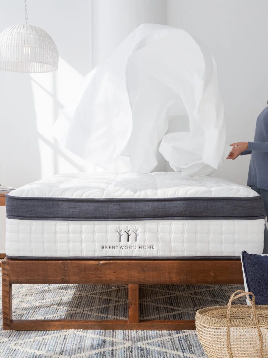 A woman stands beside a made bed, tossing a white sheet into the air in a bright, modern bedroom with a woven rug and plants.