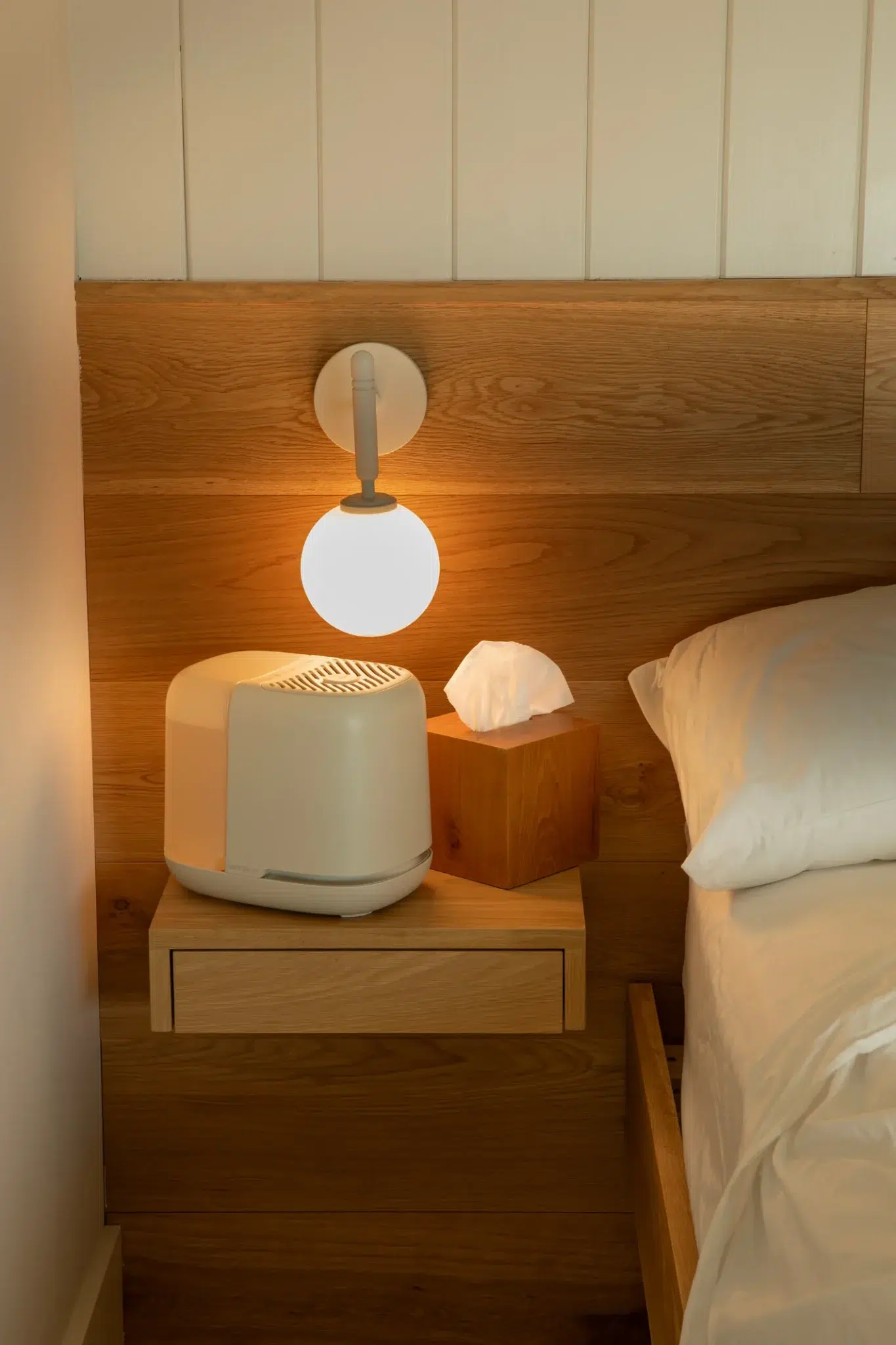 A small wall lamp, a white appliance, and a wooden tissue box sit on a wooden nightstand next to a bed with white bedding.