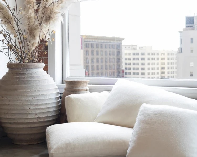 Large ceramic vase with dried grasses next to white pillows and a smaller vase by a bright window with city buildings in the background.