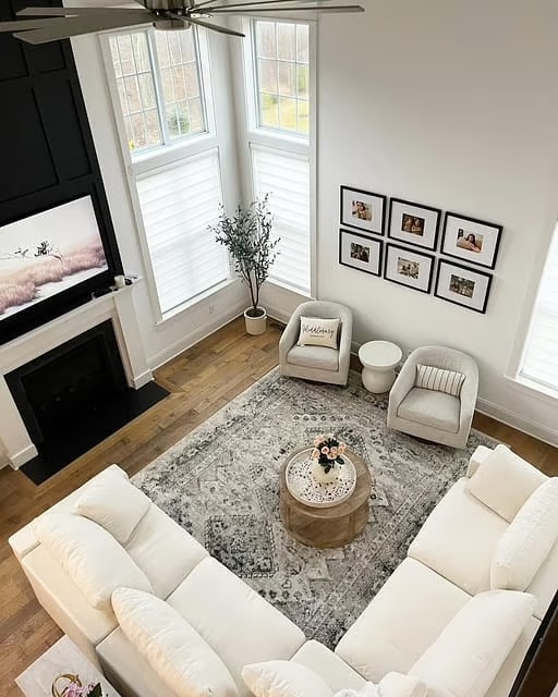 A bright living room with tall windows, a white sectional sofa, two armchairs, a round ottoman coffee table, a rug, framed photos on the wall, and a potted plant by the window.