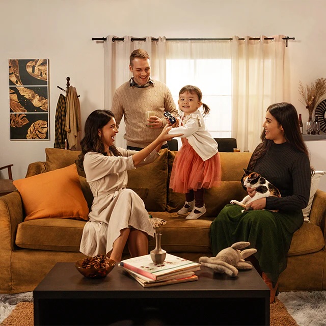Four people and a cat sit and stand in a living room. A woman hands a toy to a smiling toddler while others watch. Books, a vase, and a plush toy are on the coffee table.