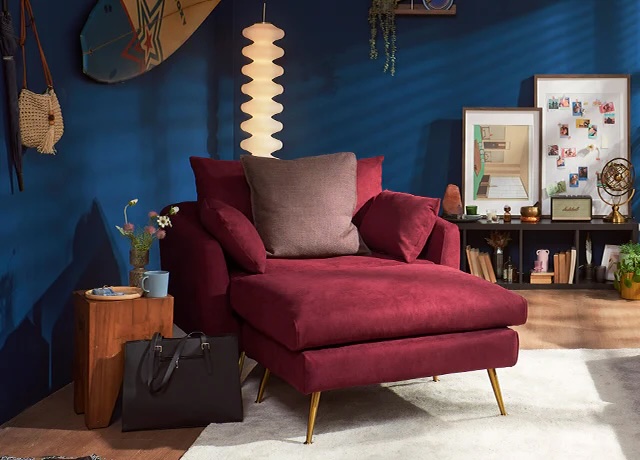 A burgundy velvet armchair with matching ottoman sits in a cozy, modern living room with blue walls, decorative items, and framed photos.