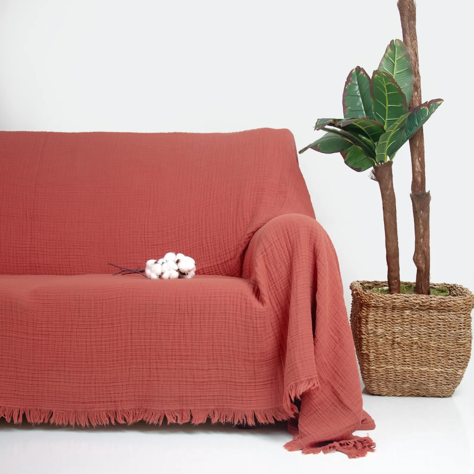 A rust-colored fringed blanket covers a couch next to a potted plant in a woven basket. A cluster of cotton rests on the couch.