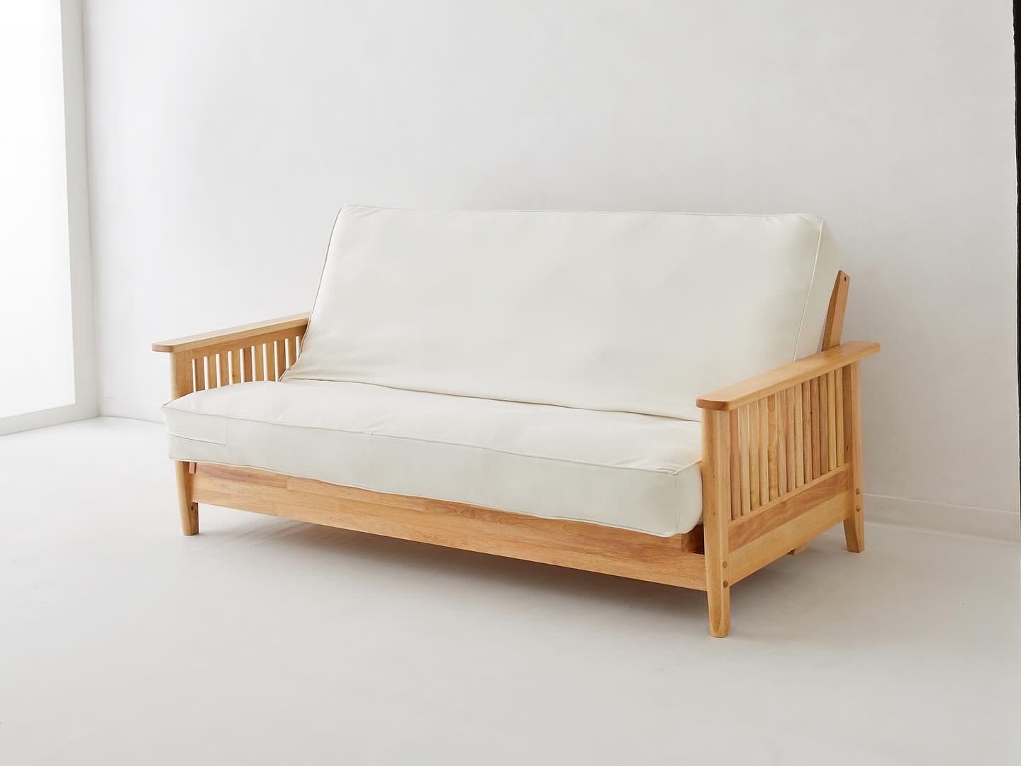 A wooden-framed sofa with white seat and back cushions sits against a plain white wall in a minimally furnished room.