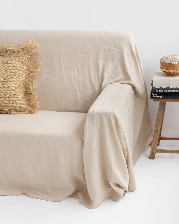 A beige linen-covered sofa with a textured cushion next to a small wooden side table holding a stack of books and a decorative item, set against a plain white background.