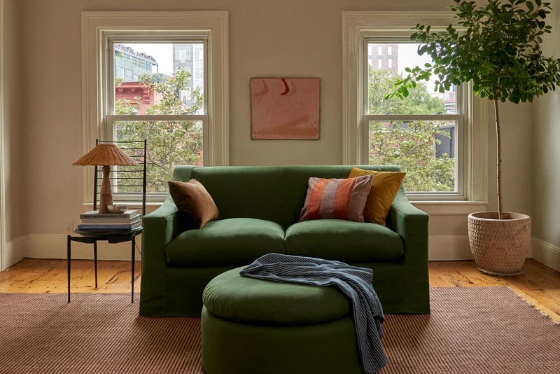 A living room with a green sofa, matching ottoman, and side table with a lamp, set between two windows; a potted tree and abstract art decorate the space.