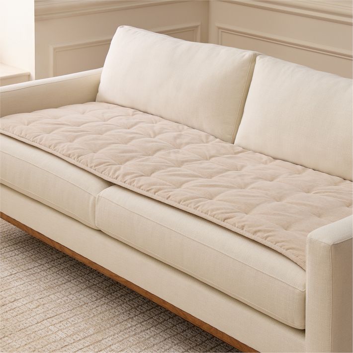 A beige quilted sofa cover is laid out neatly on a cream-colored couch in a light-toned room with a textured rug.