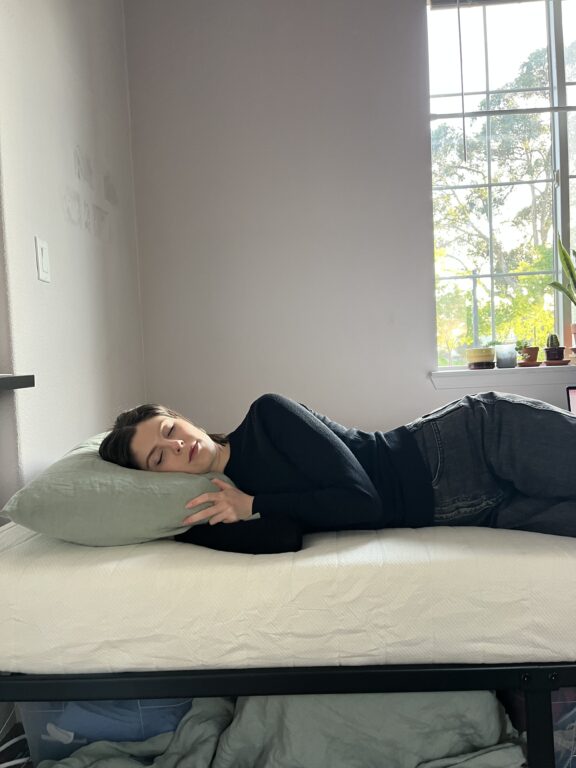 A person with short dark hair lies on their side on a bed, resting their head on a pillow, next to a window with sunlight and potted plants on the windowsill.