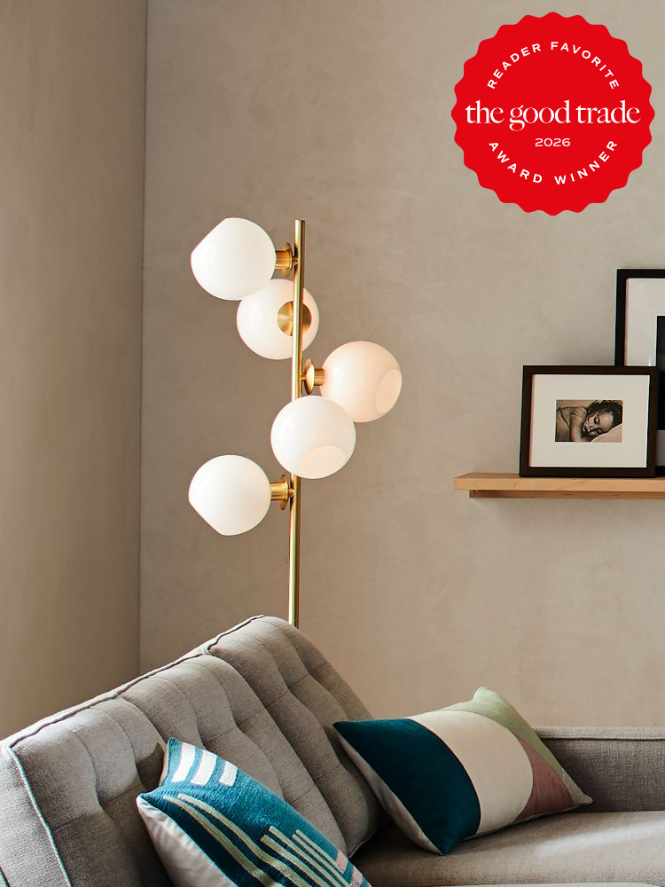 A modern floor lamp with globe-shaped bulbs stands next to a gray sofa with patterned pillows; framed photos rest on a wooden shelf above.