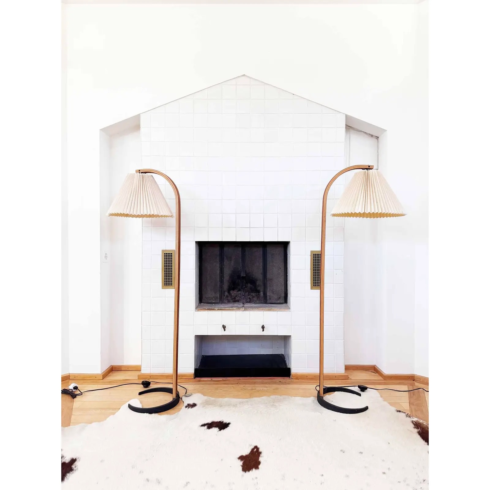 A white tiled fireplace is centered between two matching floor lamps with pleated shades. A cowhide rug covers the wooden floor in front of the fireplace.