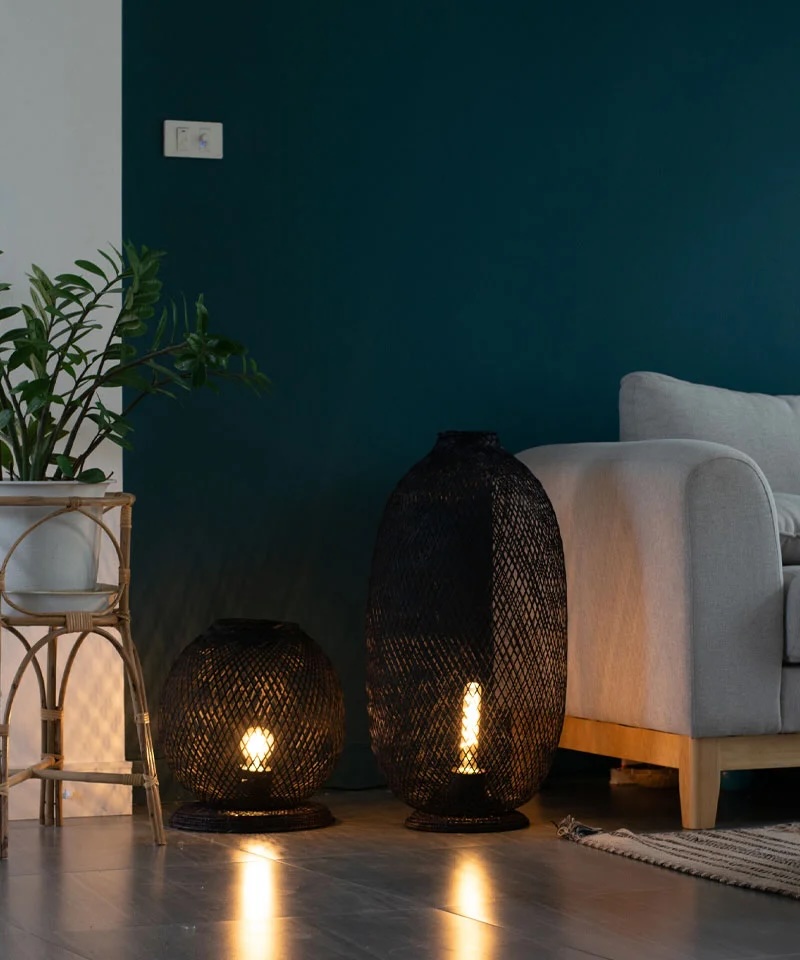 A living room corner with a gray sofa, green wall, potted plant, and two black mesh floor lamps with exposed bulbs on a wooden floor.