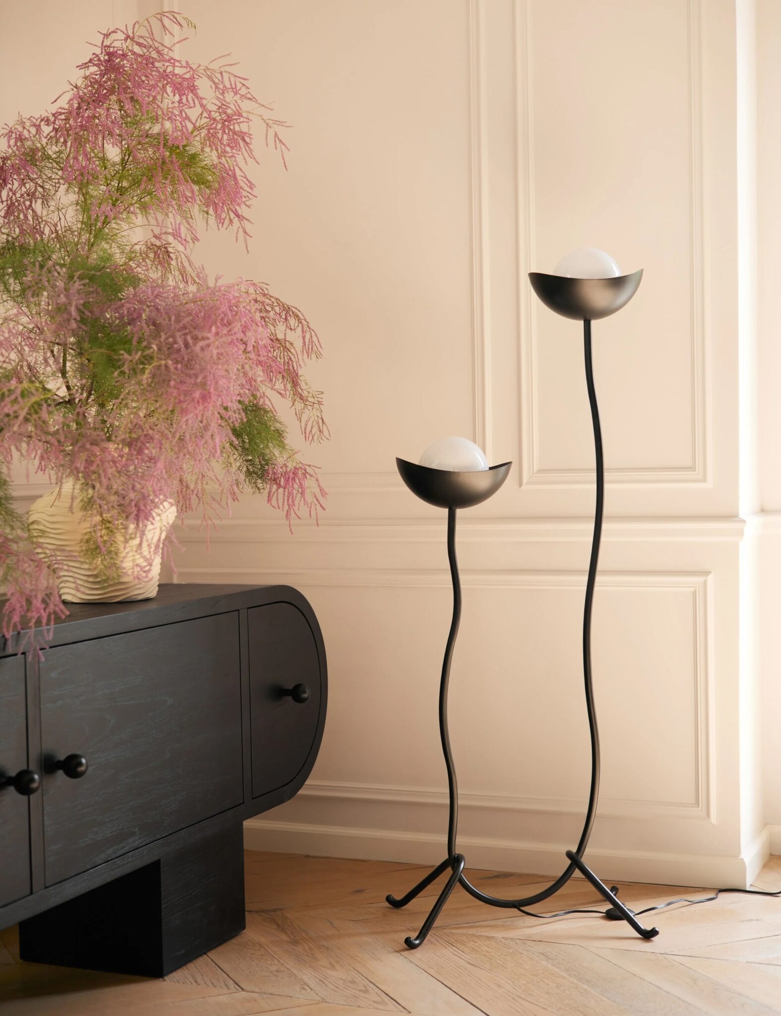 A black sideboard with round handles, a potted plant with pink feathery leaves, and two modern black floor lamps with frosted globes stand against a cream wall.