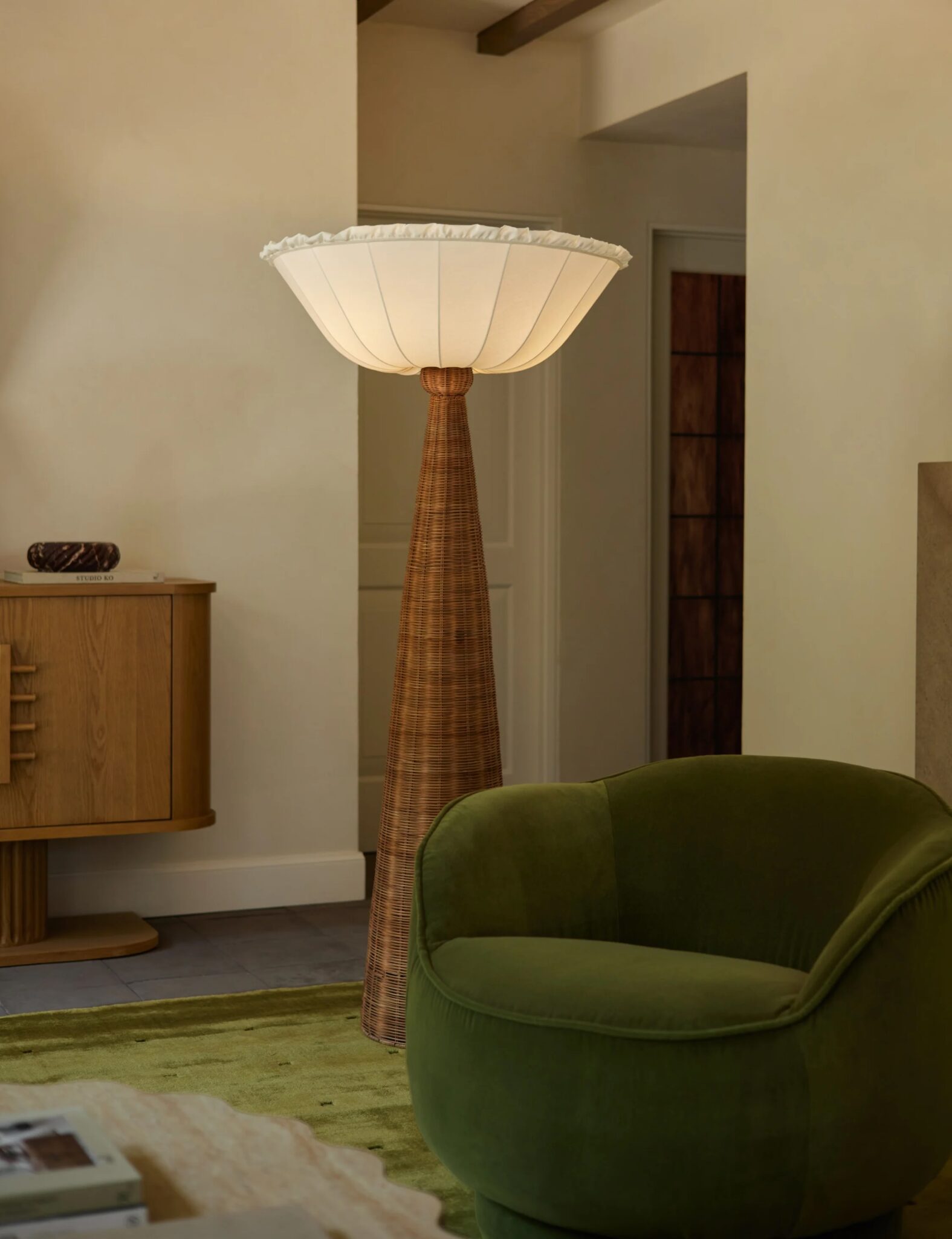 A tall floor lamp with a wide, white shade and a brown textured base stands next to a green armchair in a modern, neutral-toned room.