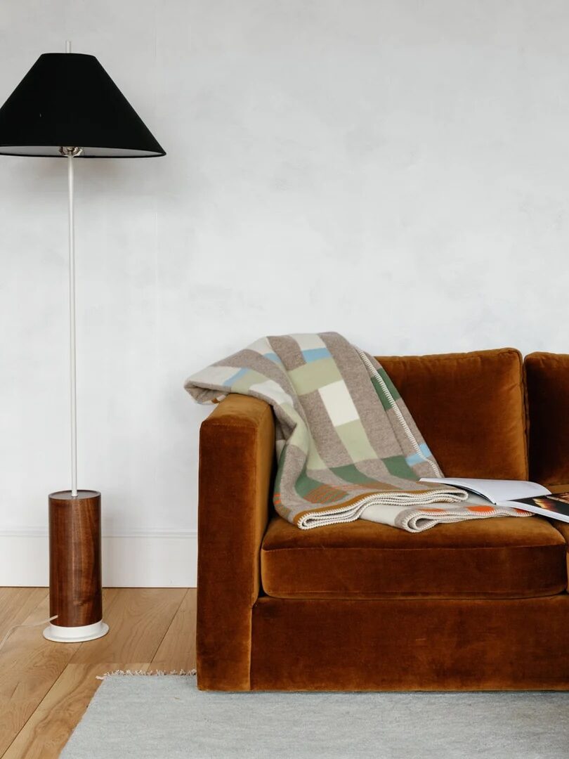 A mustard velvet sofa with a checkered blanket and open magazine sits beside a white floor lamp; a round coffee table holds a small vase.