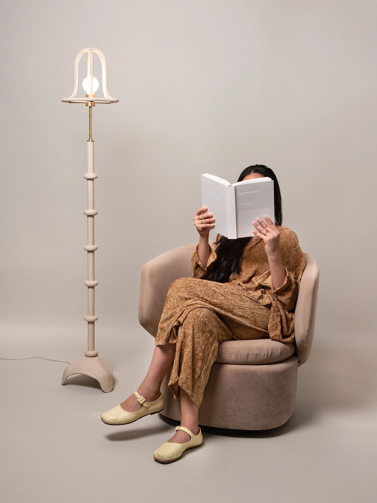 A woman in a brown outfit sits on a beige chair reading a book, with a light-colored floor lamp beside her against a plain background.