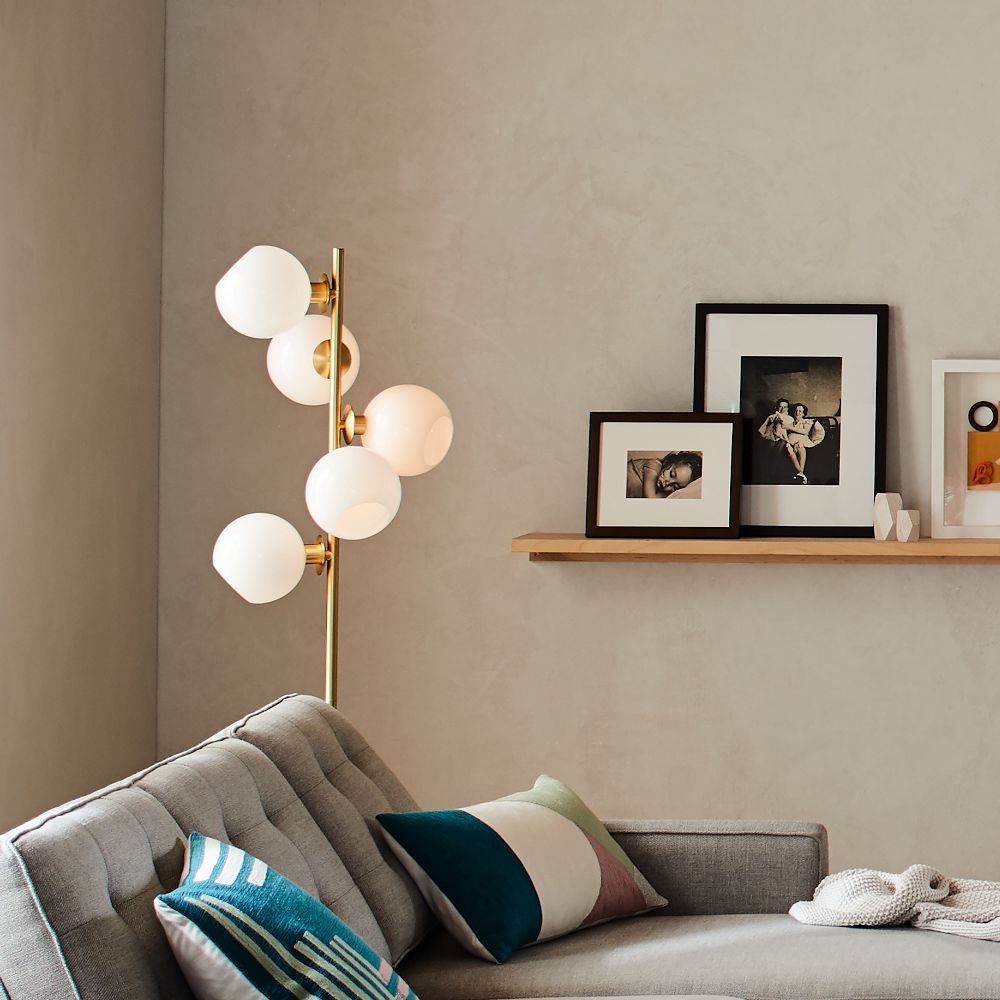 A modern living room with a gray tufted sofa, colorful pillows, a gold floor lamp with round white globes, and framed photos on a wooden shelf against a light gray wall.