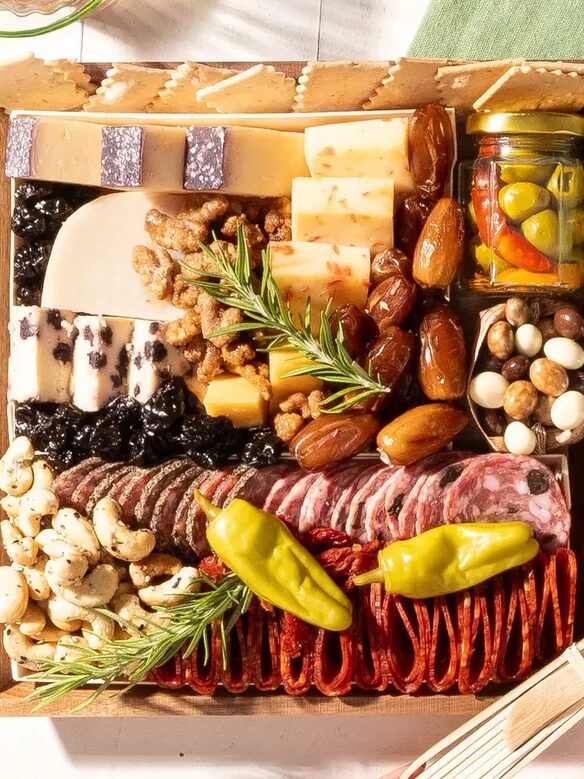A wooden box filled with assorted cheeses, meats, crackers, nuts, dried fruits, olives, pickled vegetables, and fresh rosemary sprigs.