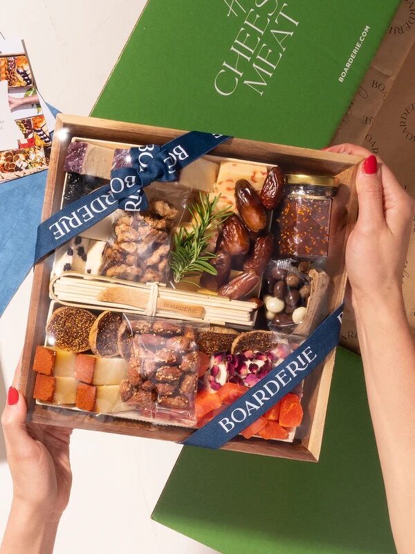 A person holds a wooden box filled with assorted cheeses, dried fruits, nuts, and snacks, labeled "Boarderie." Marketing materials and a green box are in the background.