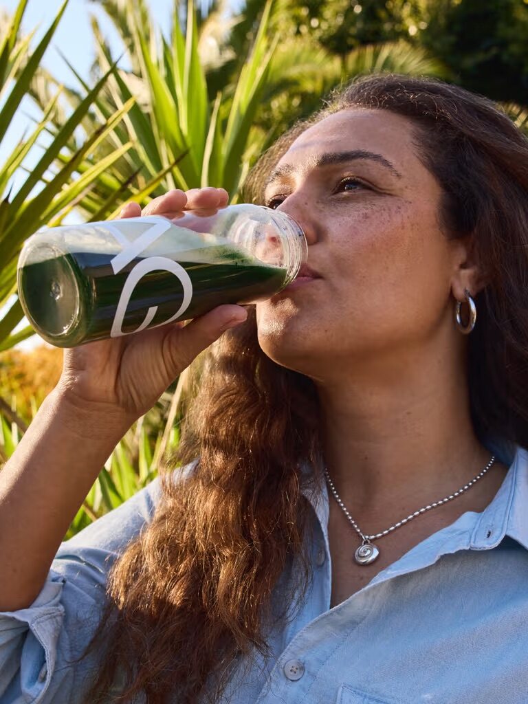 A woman with long hair drinks a green beverage from a clear bottle outdoors, surrounded by palm trees, wearing a light blue shirt and silver necklace.