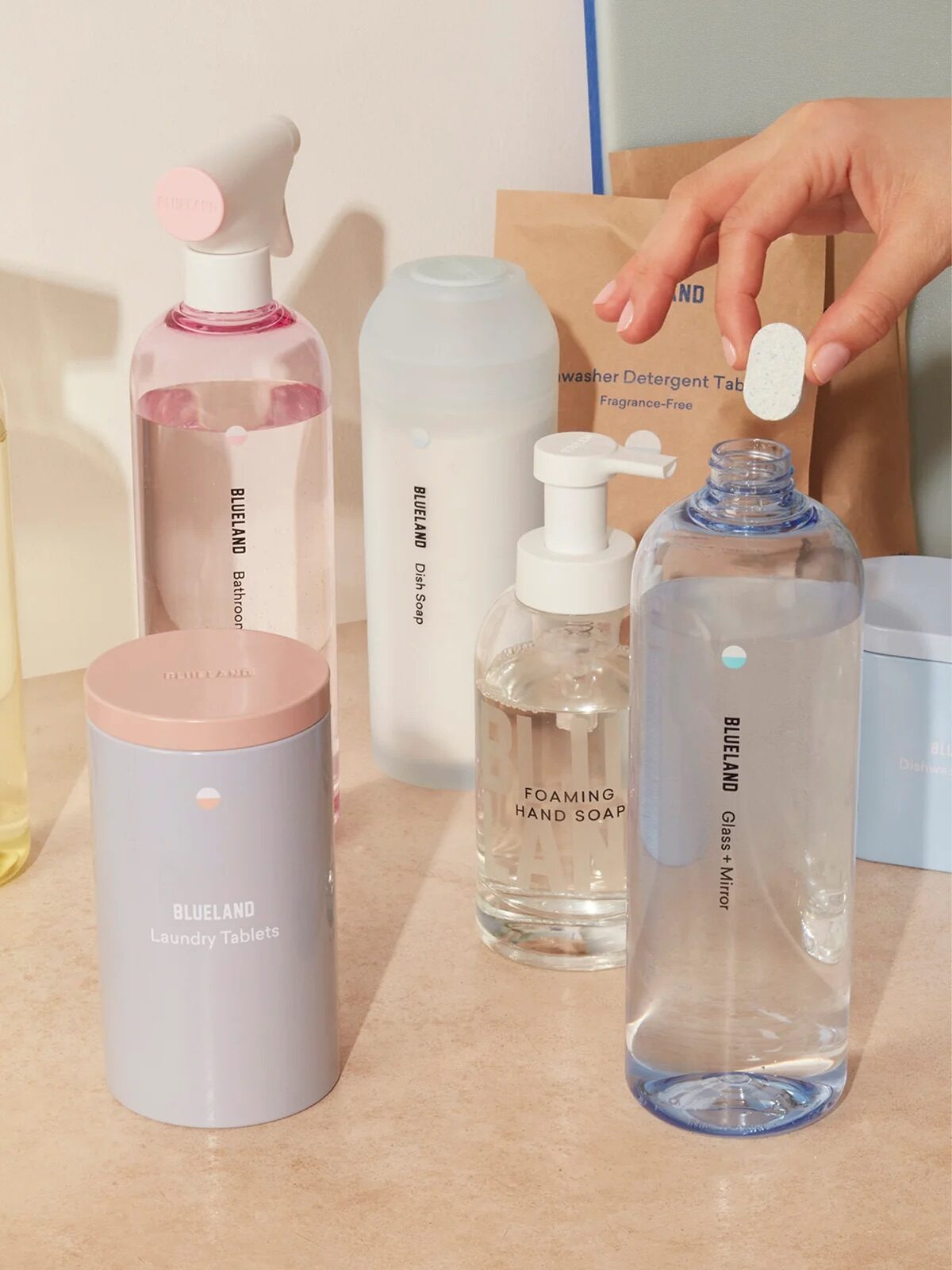 Assorted Blueland cleaning products, including spray bottles, soap dispensers, and tablets, are displayed on a counter; a hand drops a tablet into a bottle.