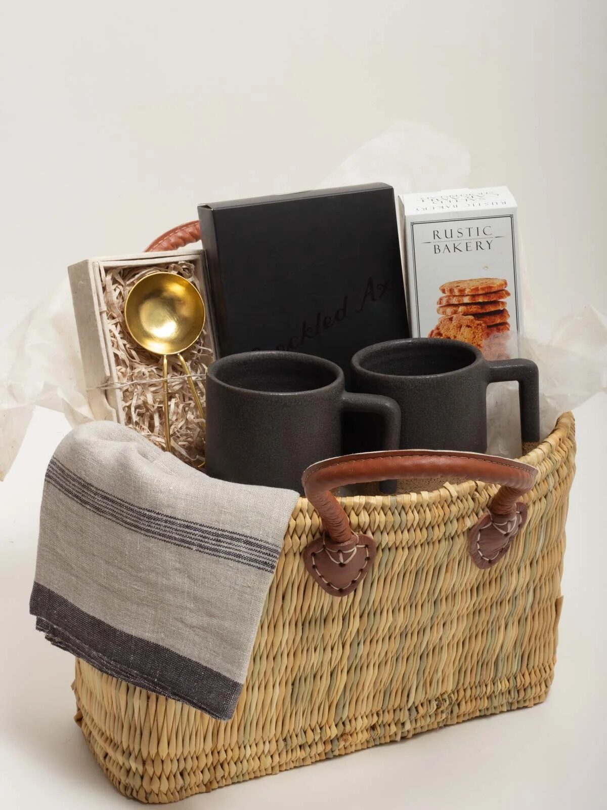 A woven basket holds two black mugs, a gray-striped cloth, a golden spoon, a black notebook, a box of crackers, and other gift items.