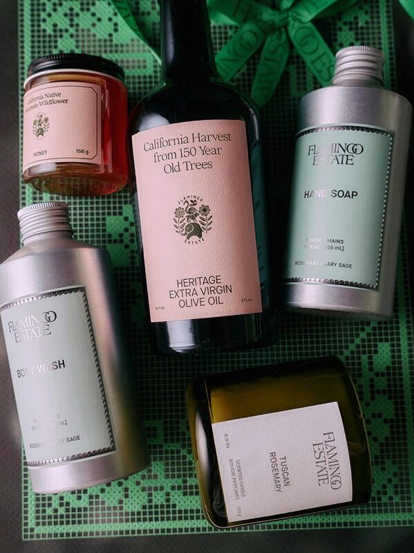 Five assorted self-care and food products, including olive oil, honey, hand soap, body wash, and a rosemary candle, are arranged on a green patterned surface.