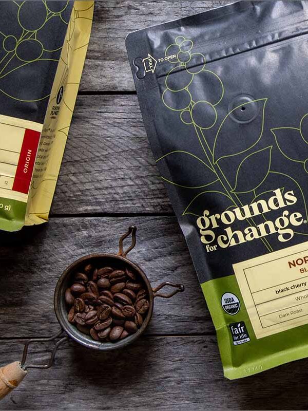 Two bags of Grounds for Change coffee, labeled Miraflor and Nordic Blend, are on a wooden surface next to a metal scoop filled with coffee beans.