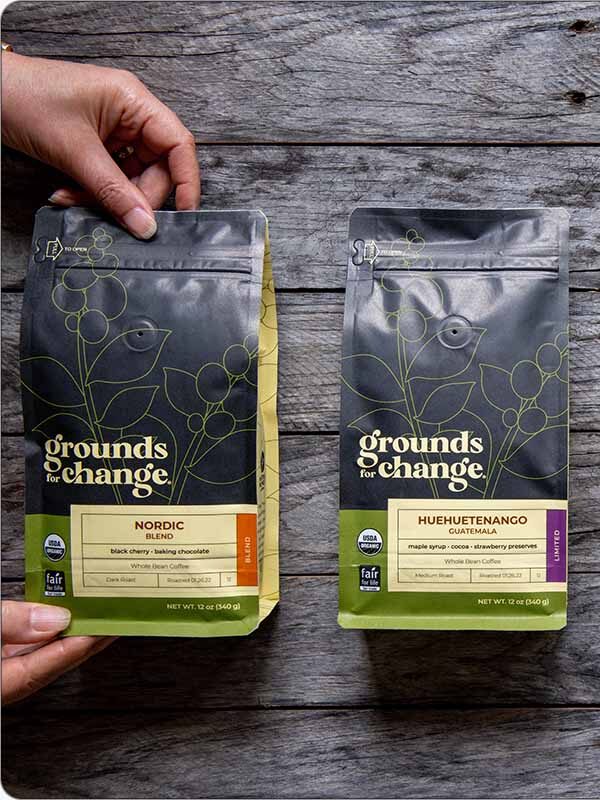 Four bags of Grounds for Change coffee—Nordic, Huehuetenango, Miraflor, and Decaf First Light—are displayed on a wooden surface. A hand holds the Nordic blend on the left.