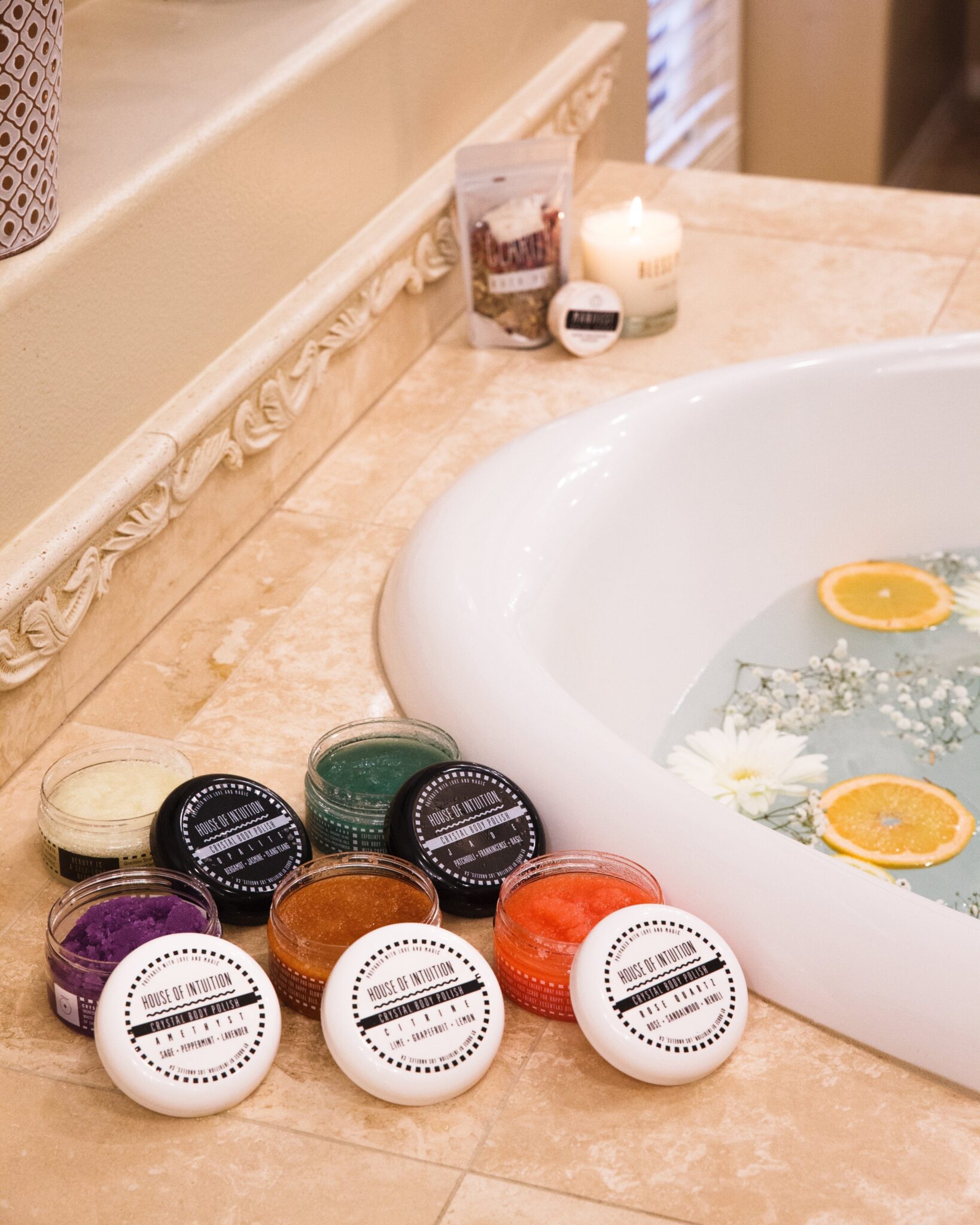 A bathroom scene with a bathtub containing water, orange slices, and flowers. Jars of bath products, a lit candle, and a small packet sit on the edge of the tub.