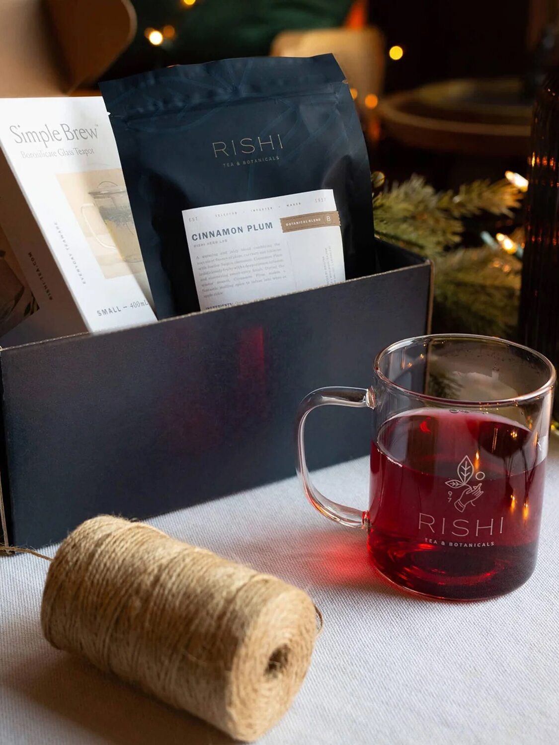 A black gift box with Rishi tea products, a glass mug of tea, a box labeled "Simple Brew," and a spool of twine on a table with festive greenery in the background.
