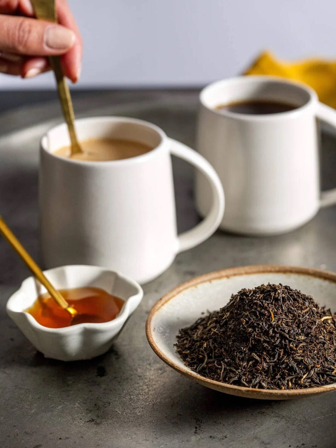 A hand stirs a mug of tea with milk beside another mug of black tea, a dish of honey with a spoon, and a plate of loose black tea leaves on a tray.