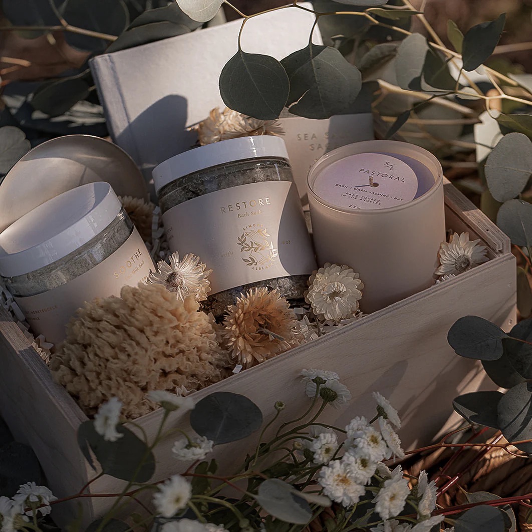 A wooden gift box contains jars of bath salts, a candle, and dried flowers, surrounded by eucalyptus leaves and white flowers.