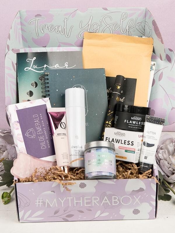 A gift box labeled “#MYTHERABOX” filled with skincare and wellness products, a notebook, and beauty items, displayed with decorative flowers.