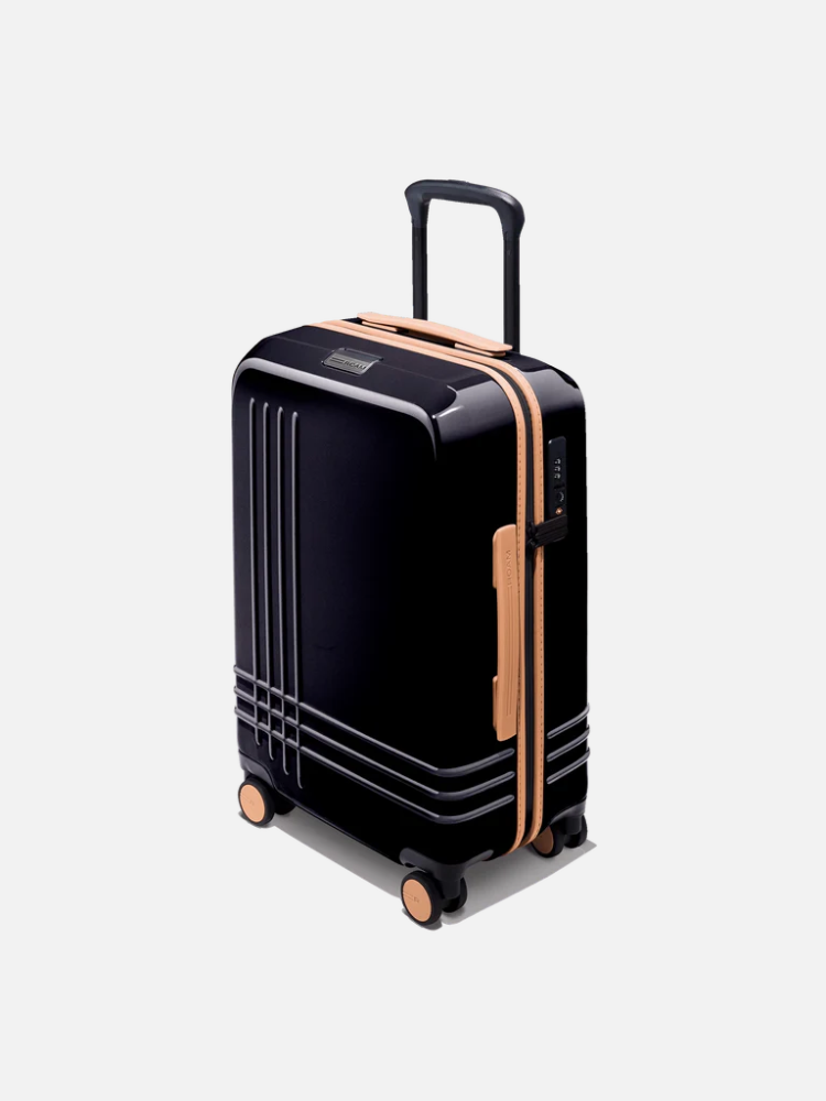 Black hard-shell suitcase with tan accents, four wheels, and a telescopic handle, standing upright on a plain light background.