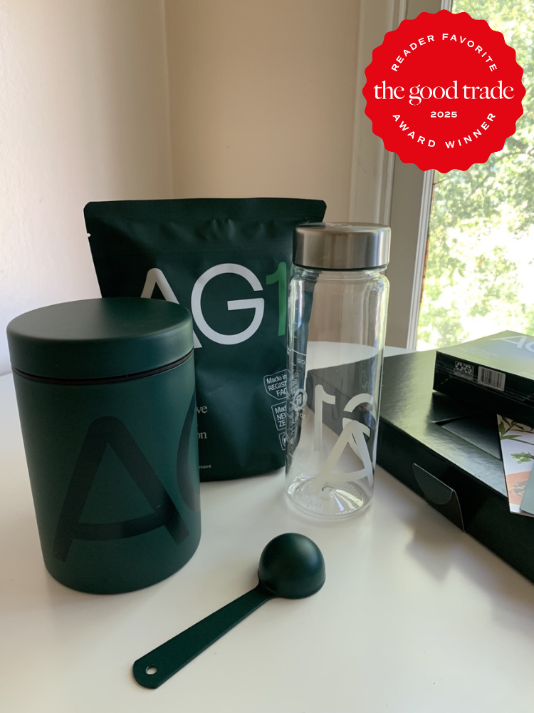 A green AG1 container, a resealable pouch, a glass bottle with a metal lid, a green scoop, and a partially open box are arranged on a tabletop.