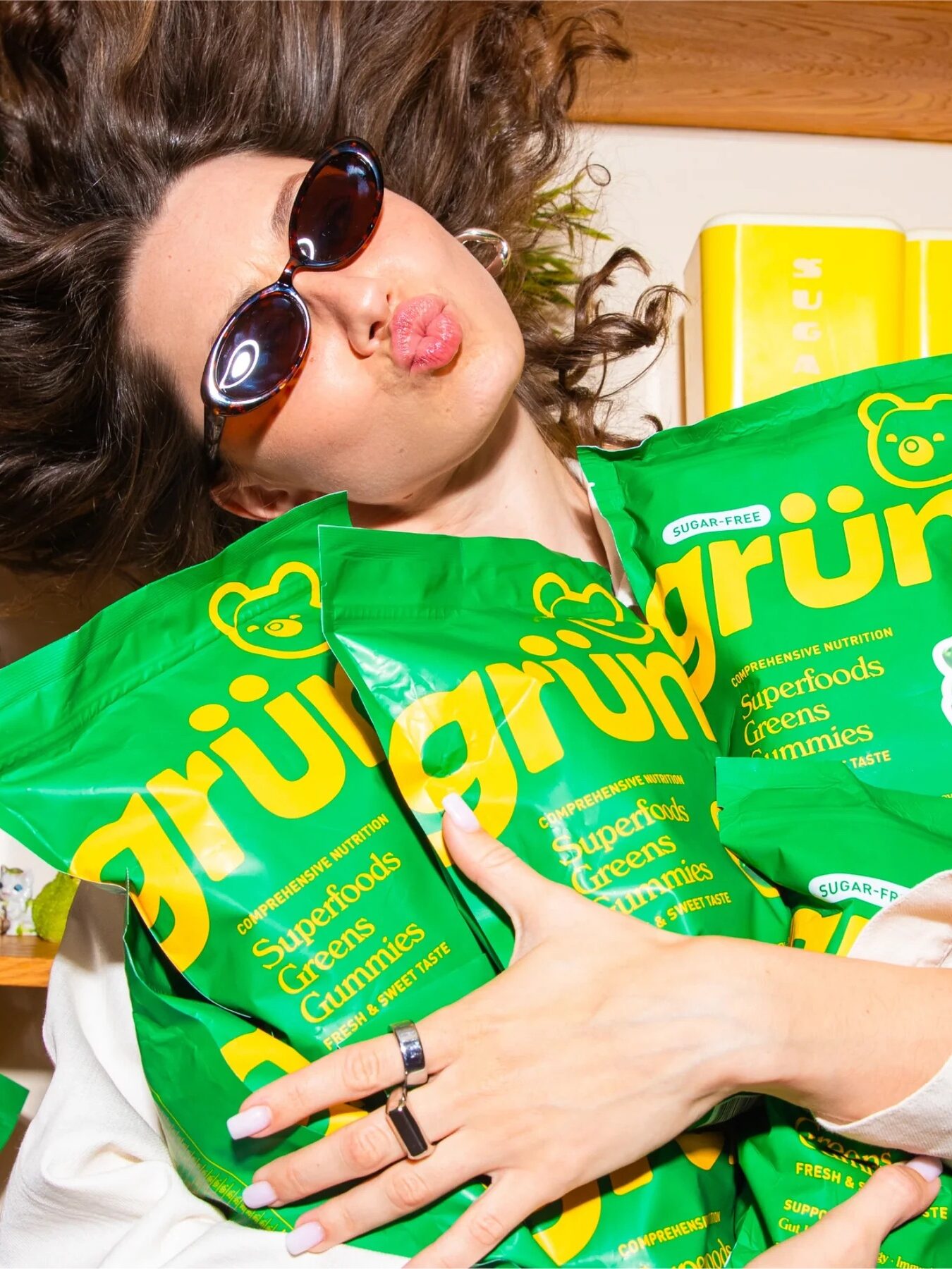 A person wearing sunglasses poses with multiple bags of Grün's Superfoods Gummies, holding them closely in front of a shelf.