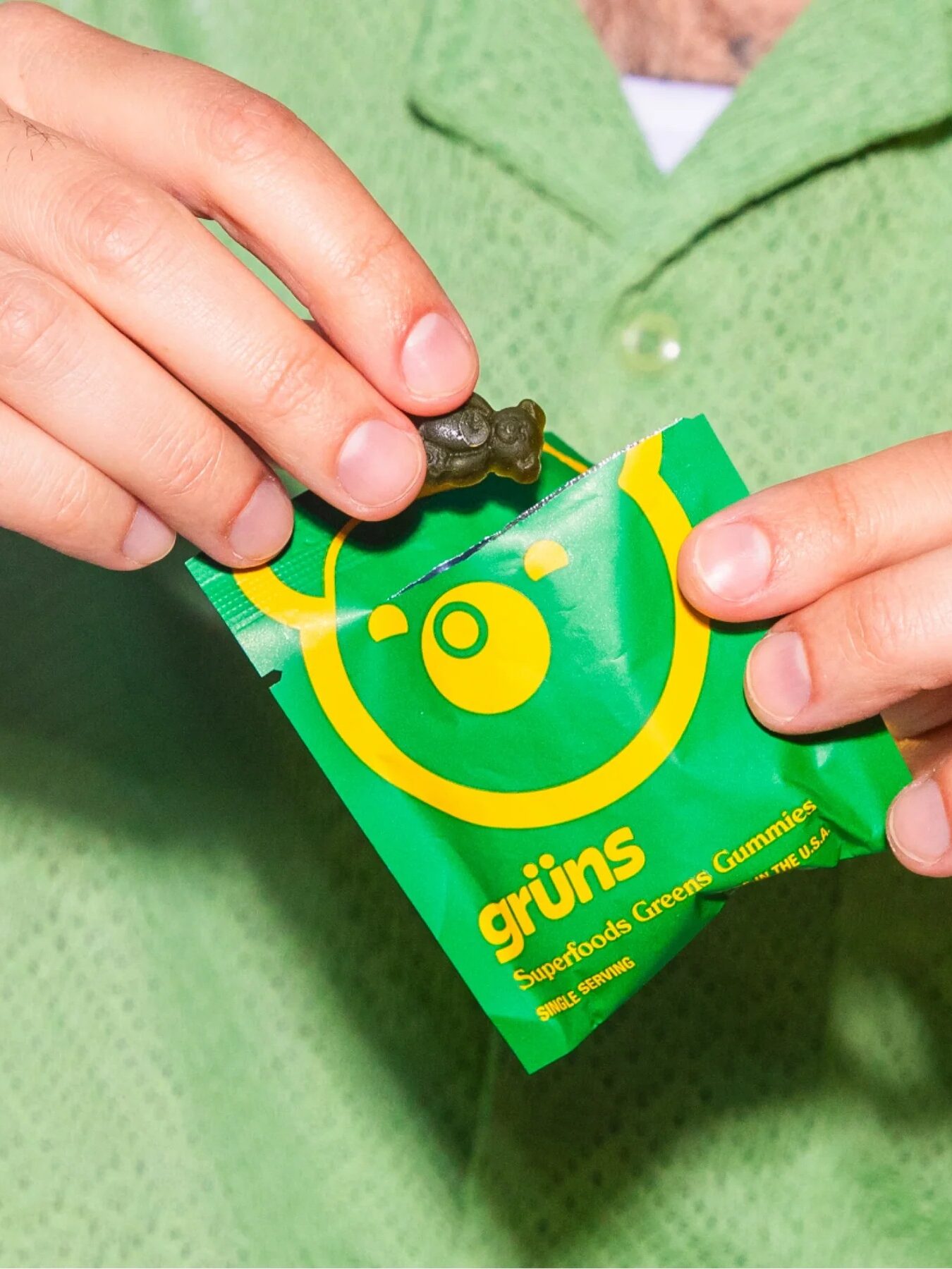 A person holds a green gummy bear above an open green and yellow packet labeled “grüns Superfoods Green Gummies.”.
