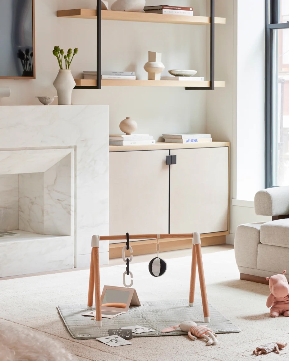A minimalist living room with a baby play gym on a rug, modern shelving, a white cabinet, and neutral-toned decor items.