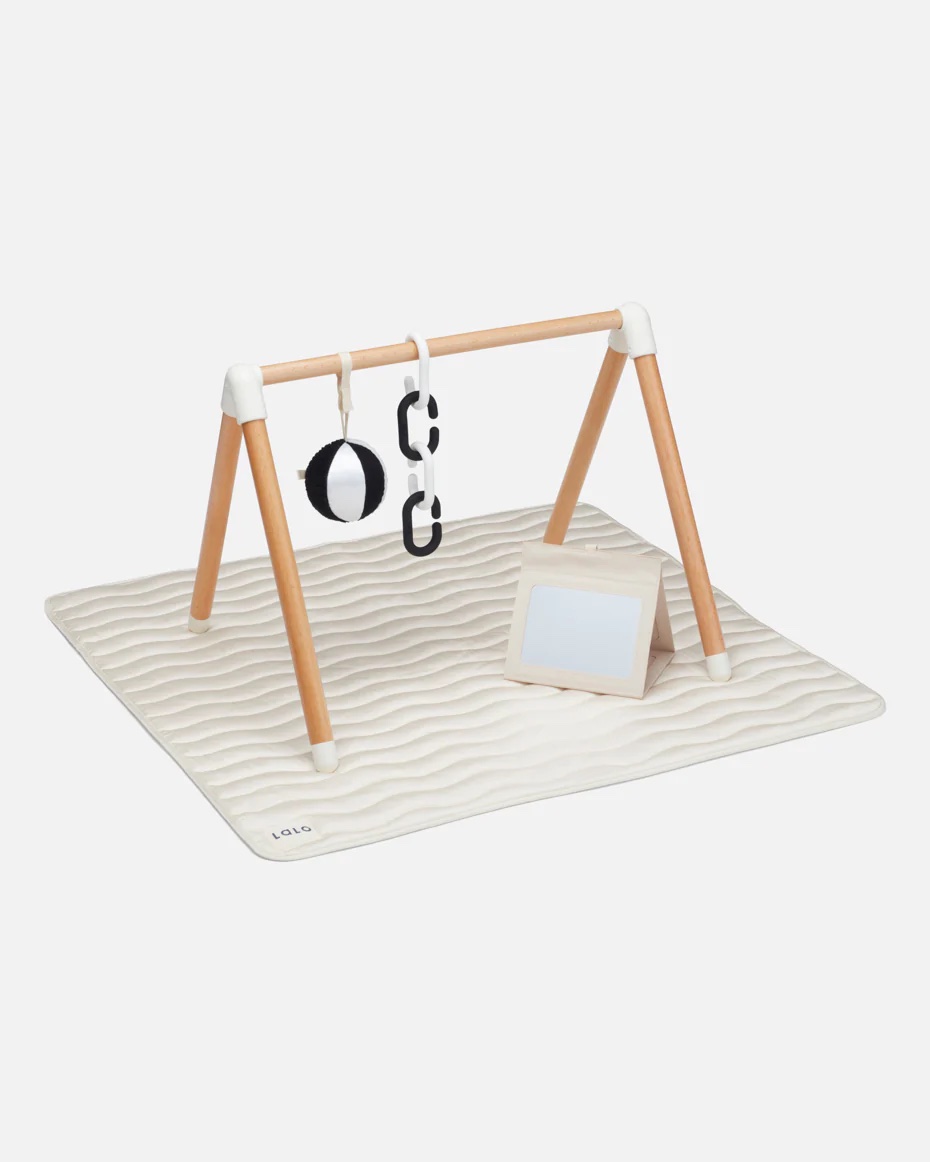 A minimalist baby play gym with a wooden frame, black and white hanging toys, a fabric mat, and an angled mirror.
