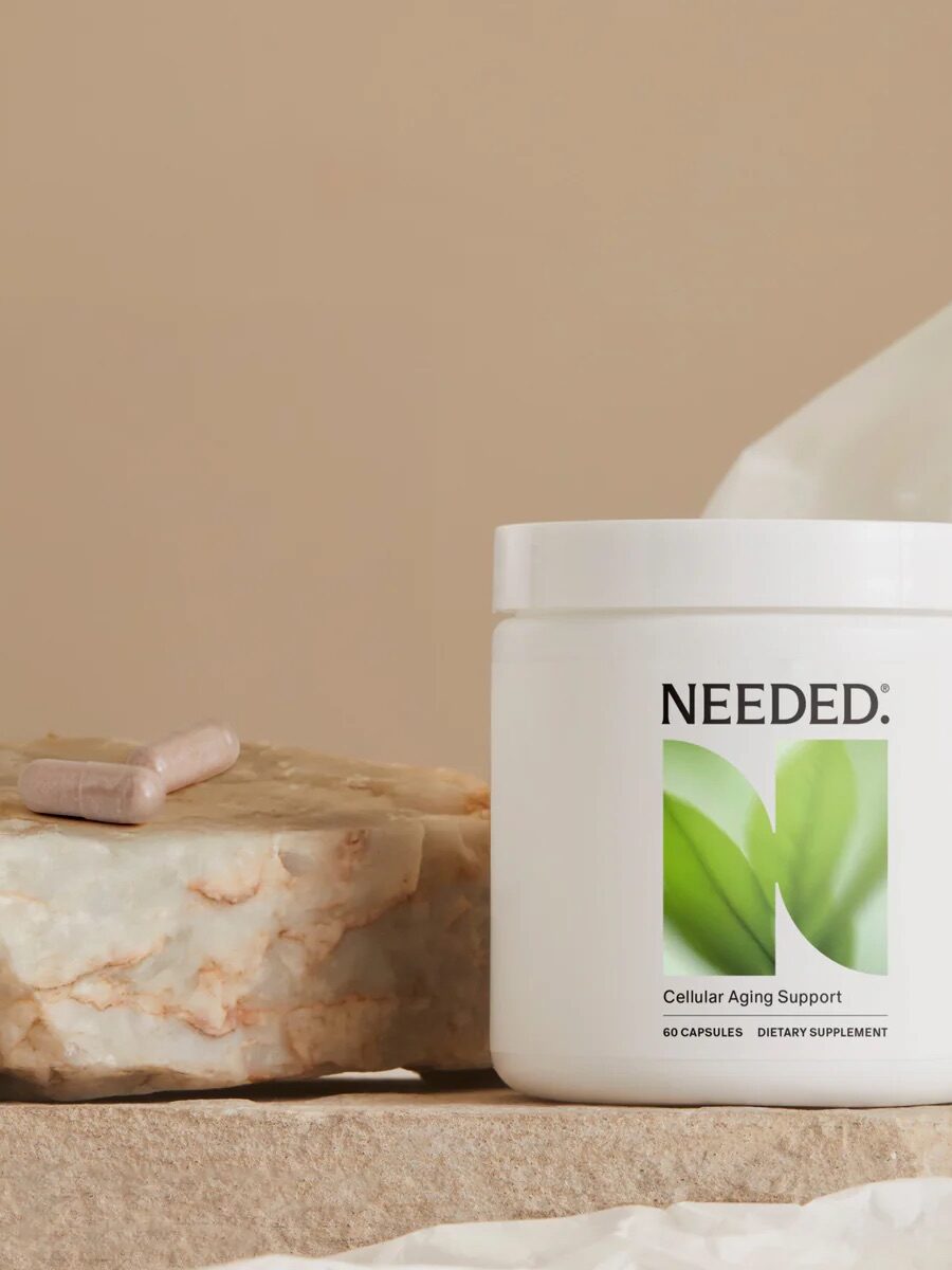 A white container labeled “Needed: Cellular Aging Support” sits on a stone surface next to two beige capsules.