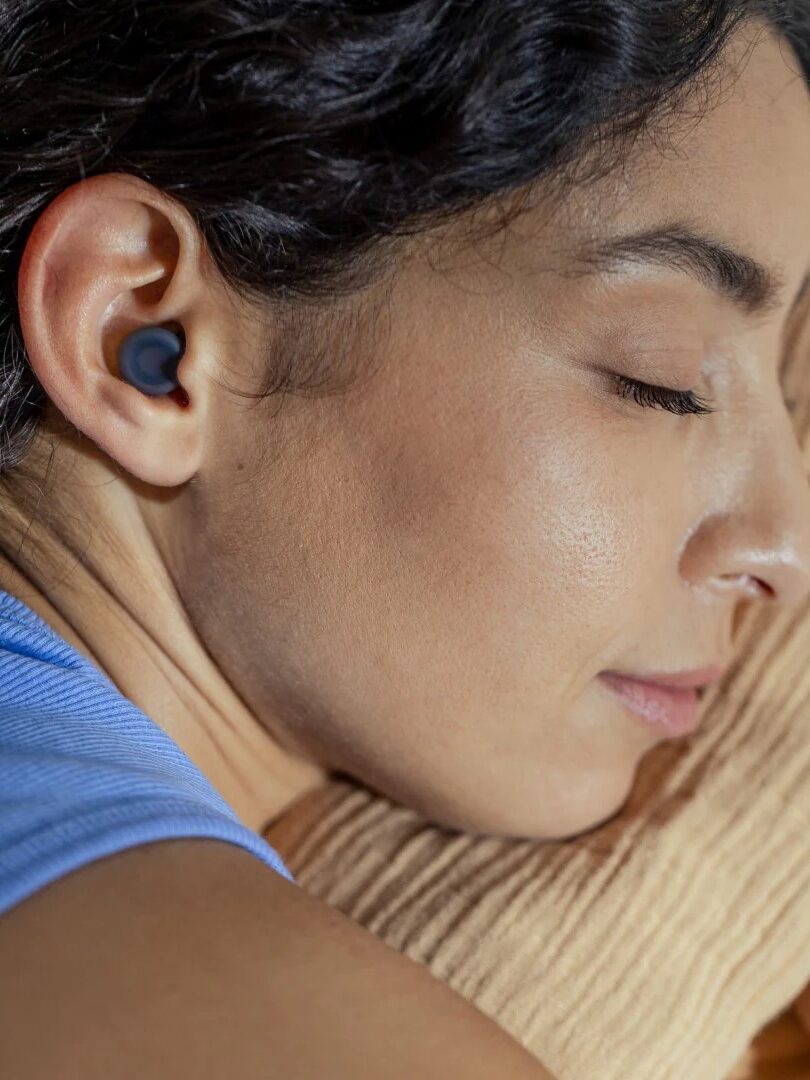 Person with dark hair lies on their side on a pillow, eyes closed, wearing a wireless earbud in one ear.