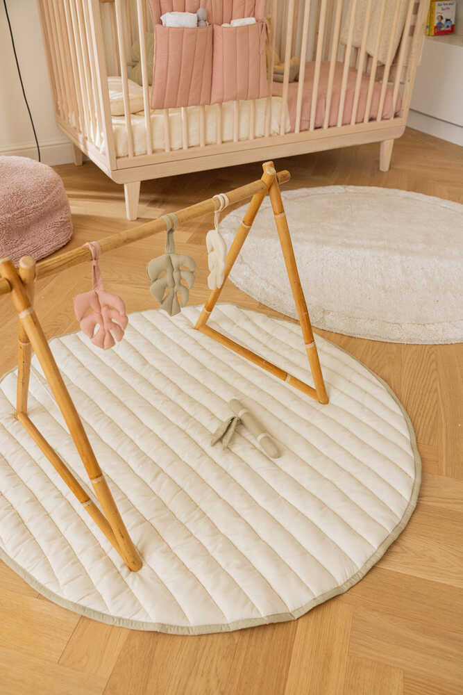 A wooden baby gym with hanging toys is set on a padded mat in a nursery with a crib, round rug, and wooden flooring.