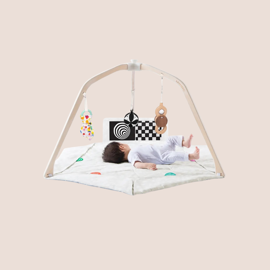 A baby lies on a play mat under an activity gym with hanging toys, including a mirror, wooden rings, and high-contrast cards.