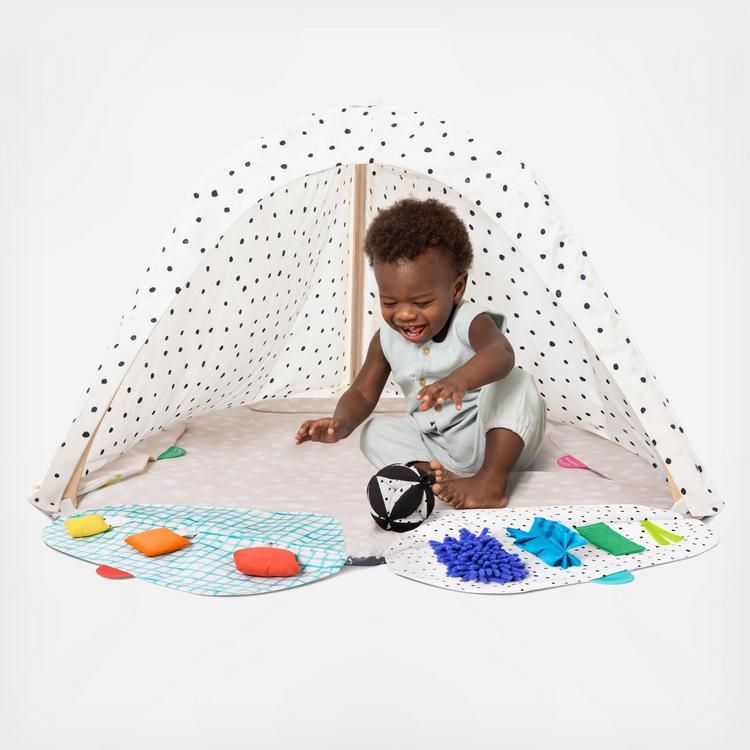 A baby sits happily inside a white, polka-dotted play tent, reaching for a black and white soft ball, surrounded by colorful textured play mats.
