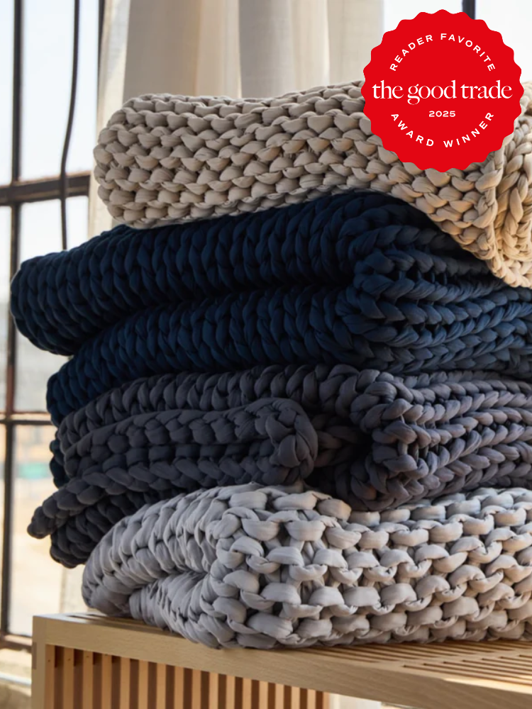 Three chunky knit blankets in cream, navy, and gray are neatly stacked on a wooden surface near a window. A red "The Good Trade 2025 Award Winner" badge is displayed in the corner.