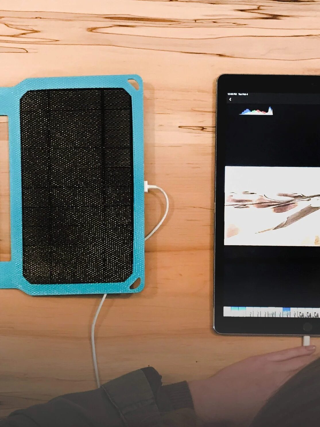 A person uses a tablet connected to a solar charger on a wooden table, with photo editing software open on the screen.
