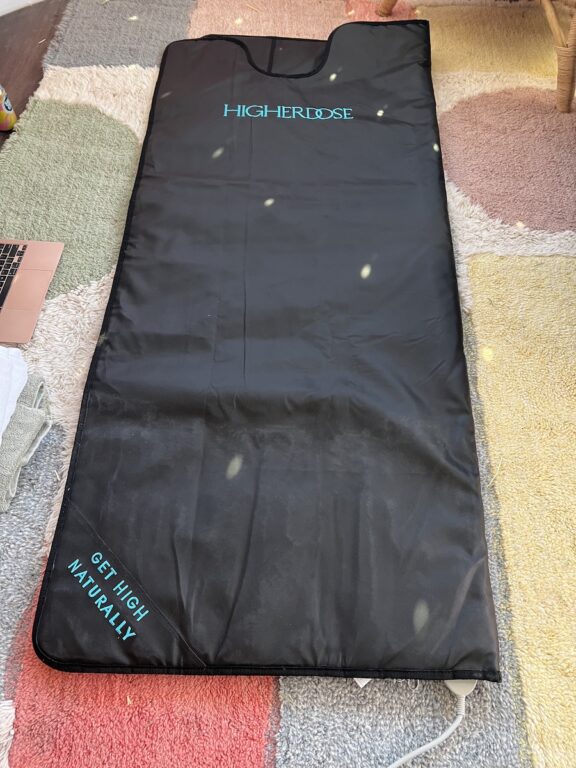 A black HigherDose infrared sauna blanket is laid out flat on a multicolored rug, with the text "GET HIGH NATURALLY" printed in blue on the bottom left corner.