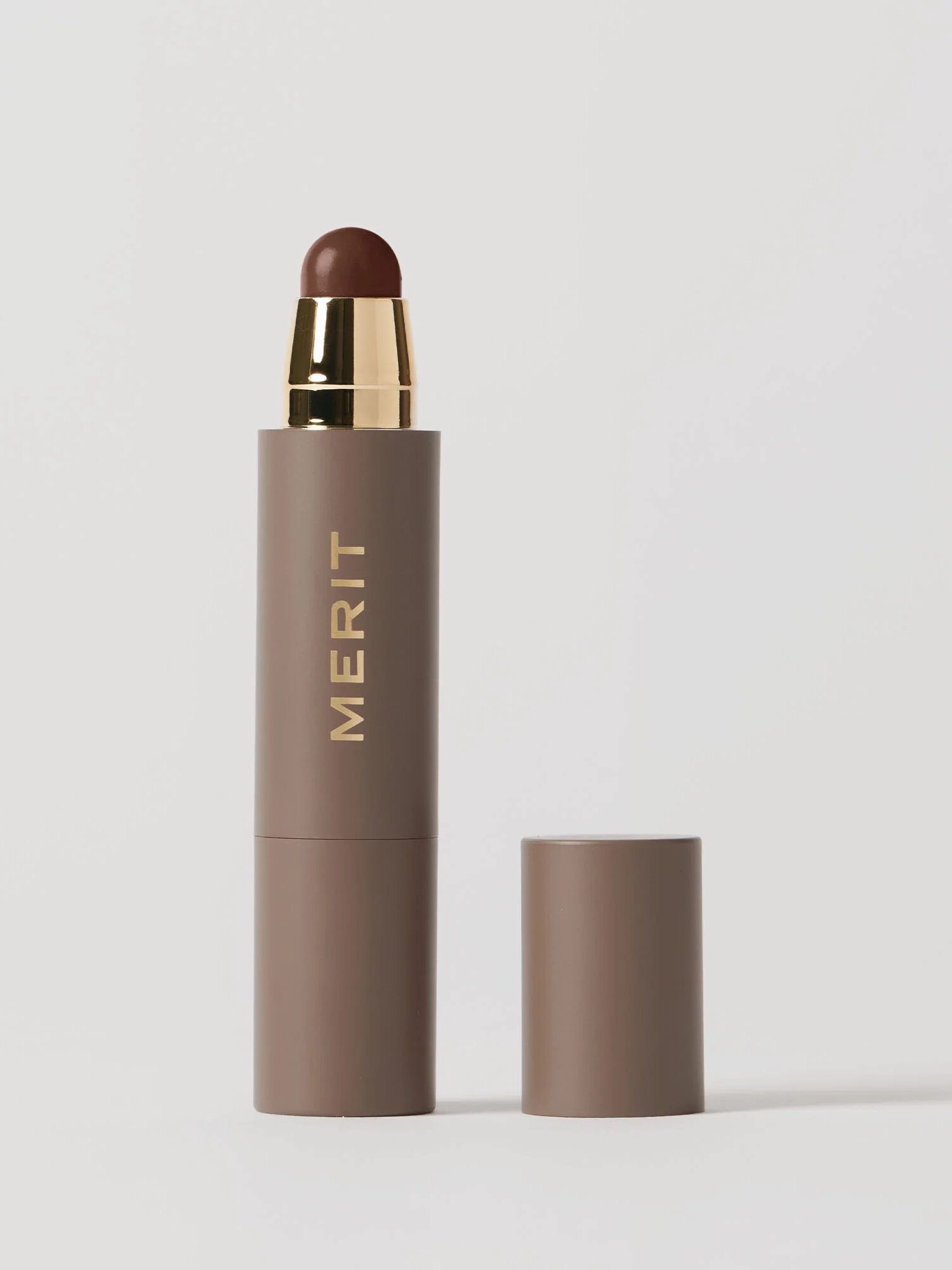 A brown stick makeup product with a gold cap and "MERIT" printed on the side, shown upright with its matching cap placed next to it on a white background.