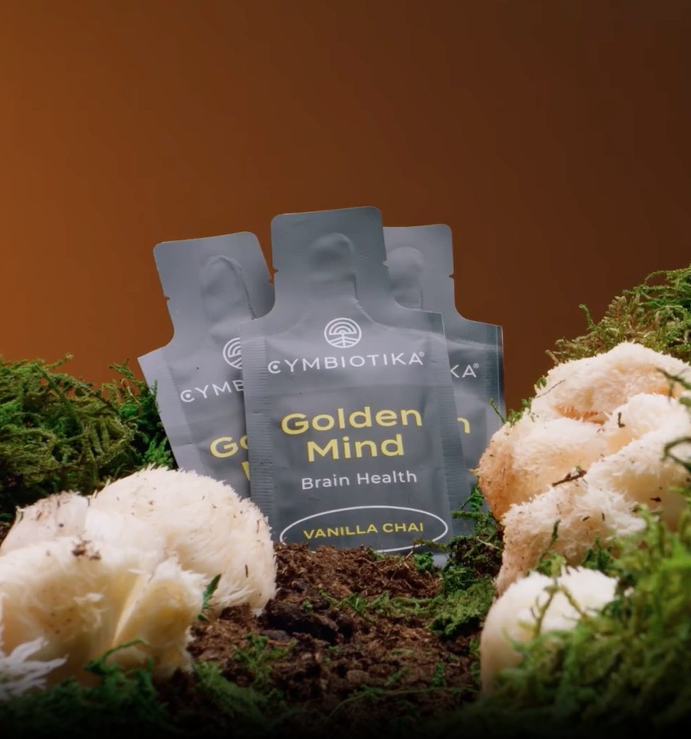 Three Cymbiotika Golden Mind Vanilla Chai brain health supplement packets stand among green moss and white mushrooms against a brown background.